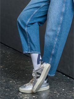 Person in blauen Jeans, weißen Socken mit schwarzem Muster und silbernen Veja-Turnschuhen, die mit einem angewinkelten Bein auf einem dunkel gefleckten Boden steht.