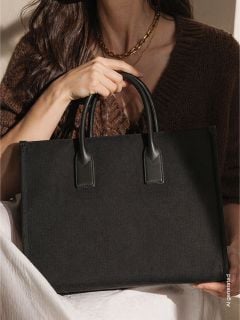 Eine Frau in einem braunen Pullover hält eine große schwarze Handtasche mit zwei Henkeln und hat eine weiße Decke auf dem Schoß.