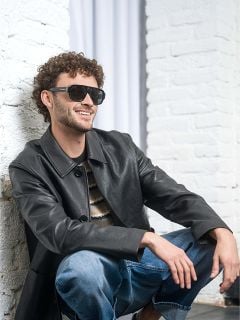 Mann mit lockigem Haar, der eine Sonnenbrille, eine schwarze Lederjacke und Jeans trägt und lächelnd vor einer weißen Backsteinmauer sitzt.
