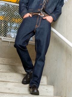 Person mit dunkler Jeans und Jacke, braunem Gürtel und Schuhen, steht auf einer Treppe und stützt sich mit der Hand auf ein Geländer. Das Gesicht ist nicht sichtbar.