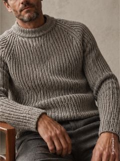 Mann, der auf einem Holzstuhl sitzt und einen dicken, gerippten grauen Pullover und eine graue Hose trägt, vor einem neutralen Hintergrund.