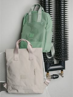 Ein hellgrüner Rucksack und eine beigefarbene Tragetasche mit Riemen stehen auf einem weißen Stuhl neben schwarzen Industrierohren.