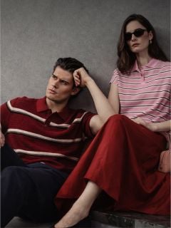Ein Mann und eine Frau sitzen an einer Wand, beide tragen gestreifte Poloshirts; der Mann trägt eine dunkle Hose, die Frau einen roten Rock und eine Sonnenbrille.