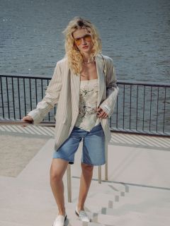 Eine Frau mit lockigem blondem Haar trägt eine Sonnenbrille, einen gestreiften Blazer, ein geblümtes Oberteil, Jeansshorts und weiße Schuhe, während sie an einer Uferpromenade mit Geländer steht.