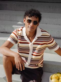 Ein Mann mit lockigem Haar und Sonnenbrille sitzt auf einer Freitreppe und trägt ein gestreiftes kurzärmeliges Hemd; neben ihm steht eine Schale mit Zitronen.