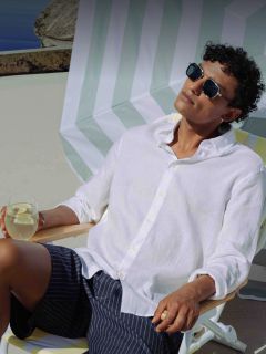 Ein Mann mit Sonnenbrille und weißem Hemd entspannt sich auf einem Liegestuhl, in der Hand ein Getränk mit Zitrone, unter einem gestreiften Sonnenschirm an einem sonnigen Tag.