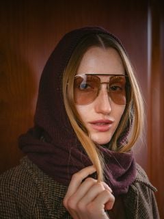 Eine Frau mit langen Haaren, die eine Pilotenbrille trägt, einen braunen Schal über dem Kopf hat und einen braunen Karomantel trägt, steht vor einer Holzwand.