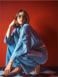 Eine Frau mit Sonnenbrille und Jeansjacke hockt auf einem schwarz-weiß karierten Boden vor einer roten Wand.