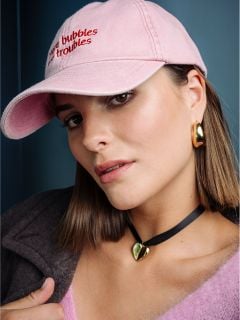 Eine Frau mit glattem braunem Haar trägt eine rosa Mütze, goldene Ohrringe, ein Herz-Halsband und ein rosa, flauschiges Oberteil und blickt vor einem blauen Hintergrund in die Kamera.