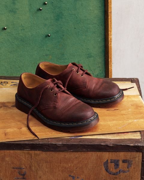 Ein Paar dunkelbraune Oxford-Schnürschuhe aus Used-Look-Leder mit gelben Nähten steht auf einer Holzfläche vor einer grünen Pinnwand.