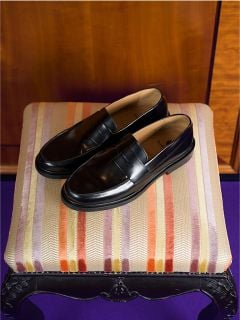 Ein Paar schwarze Leder-Loafer steht auf einem gestreiften, gepolsterten Hocker, hinter dem sich eine Holzwand befindet.