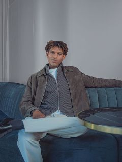 Ein Mann, der auf einem Sofa sitzt und eine graue Jacke mit Reißverschluss, ein weißes Hemd und eine weiße Hose trägt.